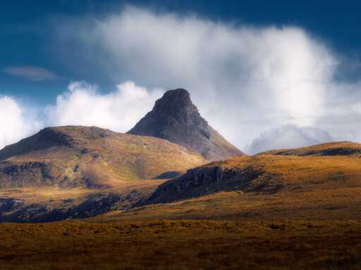 A landscape photograph of a rugged mountain rising above amber moorland under a deep blue sky. Its summit is a flat-topped, dark rock tower with stepped, layered cliffs, and its steep sides fall away in grey scree. A dense mass of white cloud billows behind the peak, and its edges glow against the blue. To the left, a long, gently sloping hillside in gold and olive tones catches the sunlight, with pale outcrops of rock along its flank. In the foreground, a broad expanse of tawny, grass-covered moor fills the lower third of the frame, with a rocky shoulder of hill running diagonally through the middle distance and a faint line of fence posts climbing towards the ridge.